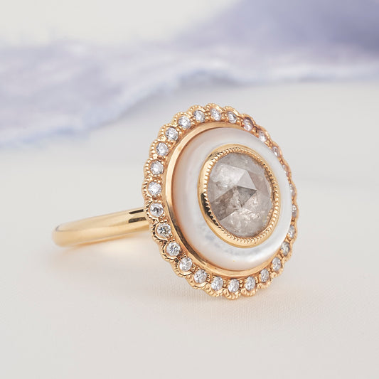 A gold ring with a large oval white mother-of-pearl inlay and a central rose-cut diamond, surrounded by a halo of small clear stones, shown on a soft light background.