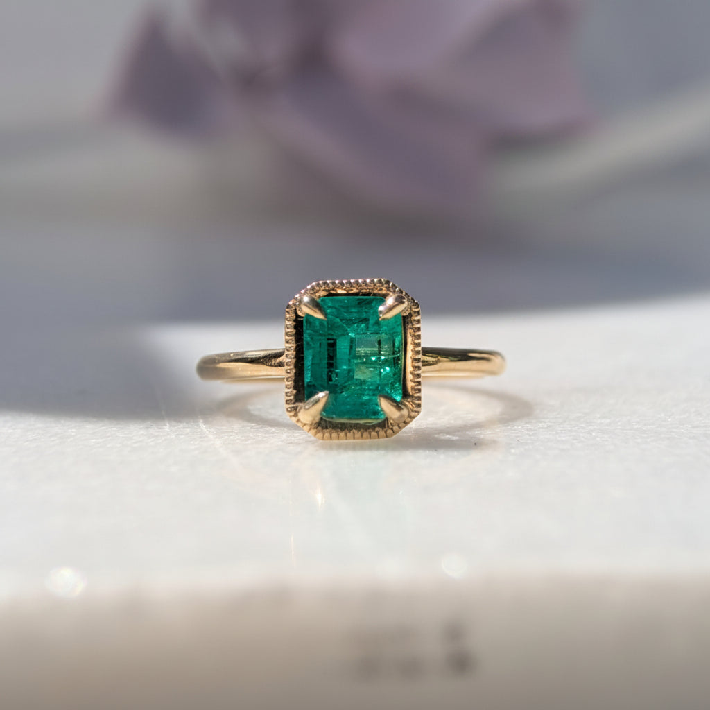 Gold ring with a green gemstone on a white surface with a blurred flower in the background