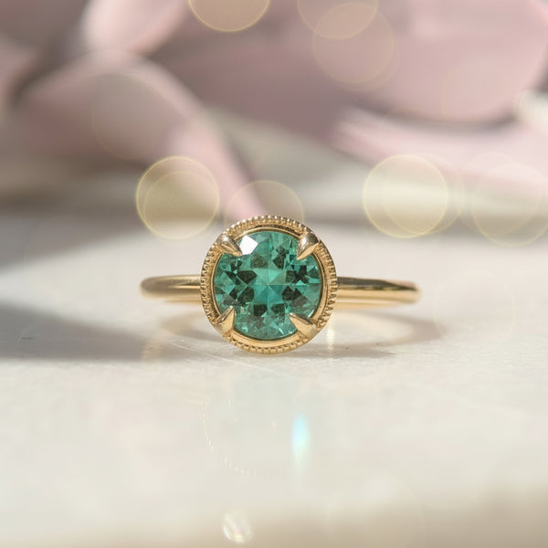 Gold ring with a green beryl gemstone on a white surface with pink flowers in the background