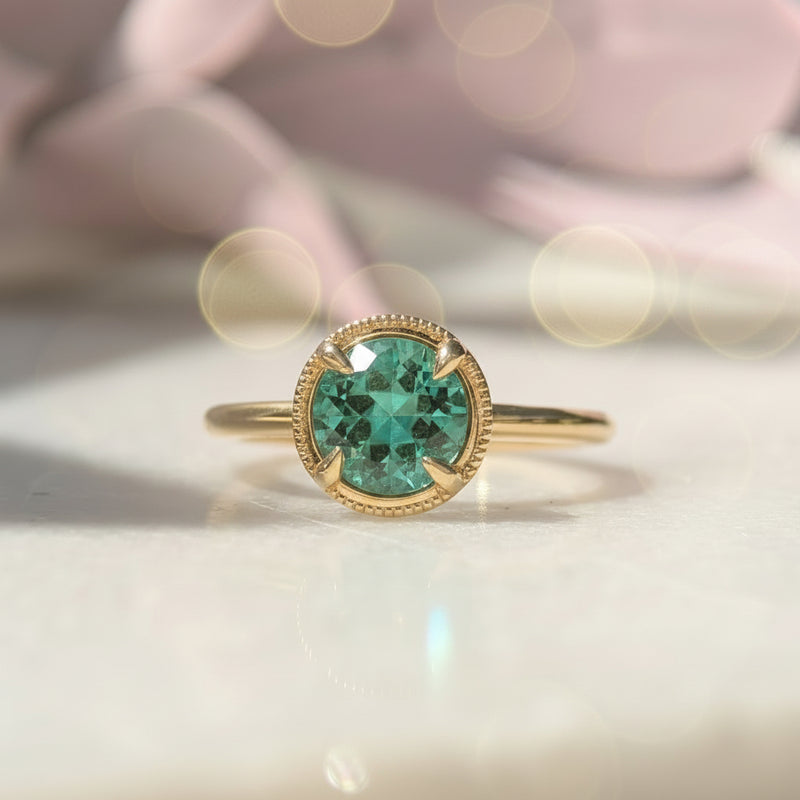 Gold ring with a green beryl gemstone on a white surface with pink flowers in the background