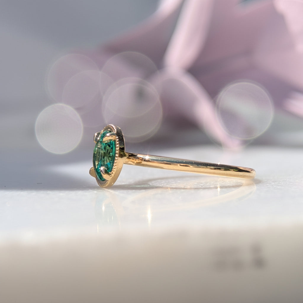 Gold ring with a green beryl gemstone on a light surface with a blurred pink flower in the background