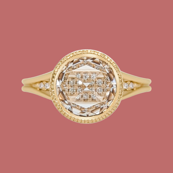 A yellow gold engagement ring featuring a round rosecut labratory created white sapphire with a split shank design and diamond accents.